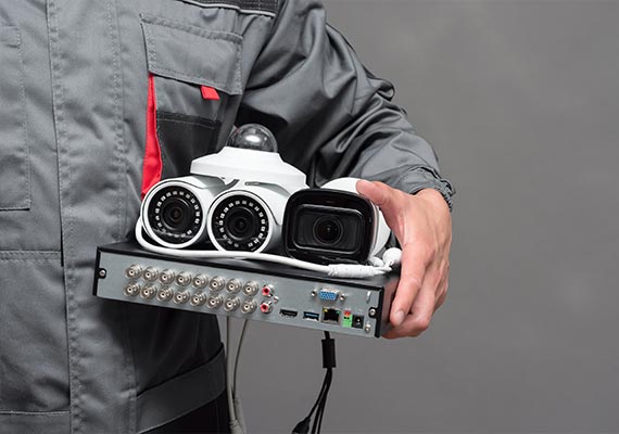 A technician in a gray uniform holds a variety of security camera equipment, including a DVR, a dome camera, and a bullet camera